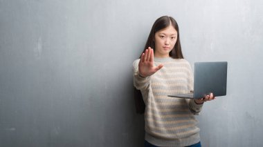 Genç Çinli kadın açık eliyle dur işareti ile ciddi ve kendine güvenen ifade, savunma hareketi yaparken bilgisayar dizüstü kullanarak gri duvar üzerinde