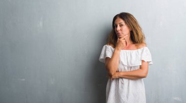 Emin çapraz kollar ve çeneme kaldırdı el ile gülümseme ile kamera seyir gri grunge duvarın üzerinden duran orta yaş İspanyol kadın. Pozitif düşünce.