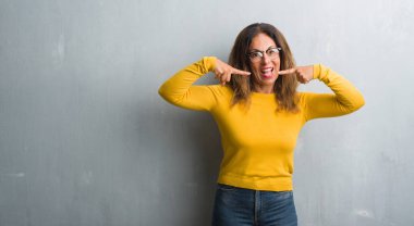 Orta yaş İspanyol kadın kendinden emin gösterilen ve işaret parmakları diş ve ağız ile gülümseyen gözlüklü gri duvar yüzünden. Sağlık kavramı.