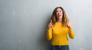 Orta yaş İspanyol kadın gözlük takan gri duvarın üzerinden hayran ve yukarıya bakıyor ve parmakları ile işaret ve silah kaldırdı şaşırttı.