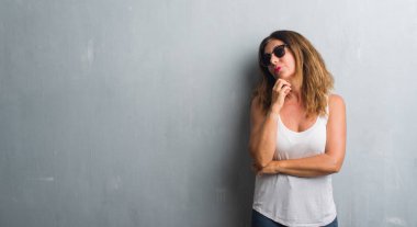 Orta yaş İspanyol kadın gri duvar çene düşünme hakkında soru, dalgın ifade üzerine el ile güneş gözlüğü takıyor. Düşünceli yüzüyle gülümsüyor. Şüphe kavramı.