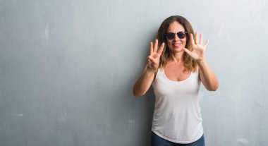 Orta yaş İspanyol kadın gösterilen ve parmakları ile işaret güneş gözlüğü takıyor gri duvar yüzünden kendinden emin ve mutlu gülümseyerek süre sekiz numara.