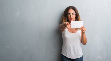 Orta yaş İspanyol kadın boş kartı kamera ve size parmak ile işaret tutan gri grunge duvarın üzerinden ayakta, el işareti, pozitif ve kendinden emin bir jest ön