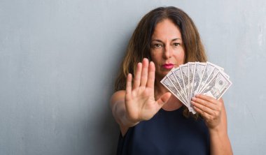 Orta yaş İspanyol kadın açık eliyle dur işareti ile ciddi ve kendine güvenen ifade, savunma jest yapıyor dolar tutan gri grunge duvarın üzerinden ayakta