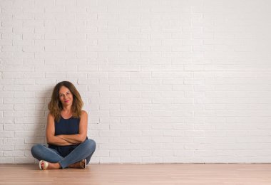 Orta yaş İspanyol kadın beyaz tuğla duvar mutlu yüz çapraz kollu kameraya bakarak gülen üzerinden yere oturma. Olumlu bir kişi.