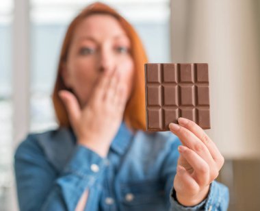 Evde çikolata bar tutan Kızıl saçlı kadın kapak ağız elle hata, korku, sessizlik içinde gizli kavramı korkmuş ifade için utanç ile şok