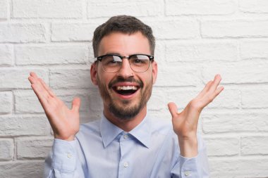 Beyaz tuğla üzerinde duran genç yetişkin iş adamı çok mutlu ve heyecanlı, kazanan ifade büyük gülümseme ile çığlık zaferini kutluyor duvar ve ellerini kaldırdı