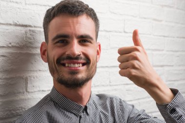Genç yetişkin adam beyaz tuğla duvardan mutlu büyük gülümseme ok işareti, başparmak ile parmak, mükemmel işareti yapıyor ayakta