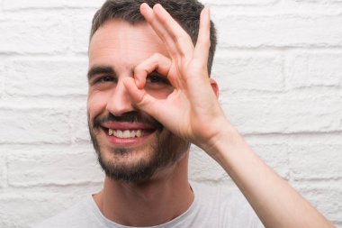 Genç yetişkin adam mutlu yüz el parmaklarının arasından seyir göz üzerinde ok işaretiyle yapıyor gülümseyen ile beyaz tuğla duvarı ayakta