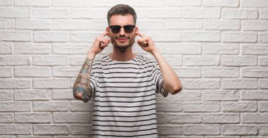 Genç yetişkin adam beyaz tuğla duvar kulakları parmakları ile yüksek sesle müzik gürültü rahatsız ifade ile kaplama üzerinde duran güneş gözlüğü takıyor. Sağır kavramı.