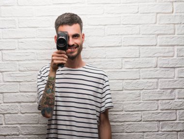 Genç adam Holding video bir gülen surat konumu olan beyaz tuğla duvar üzerinde duran ve diş gösterilen kendine güvenen bir gülümsemeyle gülümseyerek kamera çekimi