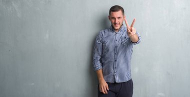 Beyaz delikanlı zafer işareti yapıyor parmak gösterilen kamera seyir gülümseyen gri grunge duvarın üzerinden. İki numaralı.