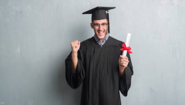 Duygu tezahürat Genç beyaz adam gurur çığlık ve zafer ve başarı çok heyecanlı, kutluyor derecesi tutan lisansüstü üniforması gri grunge duvarın üzerinden