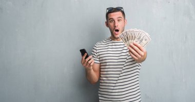 Dolar ve smarphone korkmuş şok bir sürpriz yüz, korkuyor ve heyecanlı korku ifadesi ile tutarak gri grunge duvarın üzerinden beyaz delikanlı