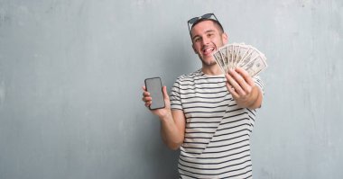 Beyaz delikanlı dolar ve smarphone dil yapışkanlık dışarı komik bir ifadeyle mutlu tutarak gri grunge duvarın üzerinden. Duygu kavramı.