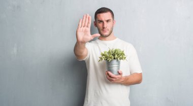 Bitki pot ile tutarak gri grunge duvarın üzerinden beyaz delikanlı dur işareti ile ciddi ve kendine güvenen ifade, savunma hareketi yaparken el açmak