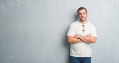 Beyaz delikanlı yüz çapraz kolları üzerinde güneş gözlüğü şüpheci ve sinir, onaylamayan ifade giyen gri grunge duvarın üzerinden. Negatif kişi.