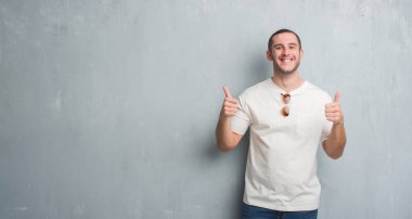 El ile olumlu hareketi yaparken güneş gözlüğü başarı işareti giyiyor gri grunge duvarın üzerinden beyaz delikanlı başparmak gülümseyen ve mutlu. Neşeli bir ifadeyle, kazanan jest kameraya bakıyor.