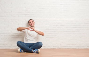 Kalp simgesi ve elleriyle şekil gösterilen aşık gülümseyen beyaz tuğla duvarın üzerinden katta oturan beyaz delikanlı. Romantik kavramı.