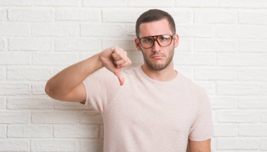 Genç beyaz adam kızgın yüzü, eksi işareti aşağı antipati başparmak ile gösterilen, ret kavramı ile gözlüklü beyaz tuğla duvarın üzerinden ayakta