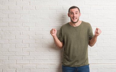 Genç beyaz adam ayakta beyaz tuğla duvar kutluyor üzerinde şaşırttı ve başarı için kaldırdı kollarını açarak hayret ve gözleri açık. Kazanan kavramı.