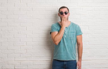 Genç beyaz adam güneş gözlüğü kapak ağız elle hata, korku, sessizlik içinde gizli kavramı korkmuş ifade için utanç ile şok giyen beyaz tuğla duvarın üzerinden ayakta