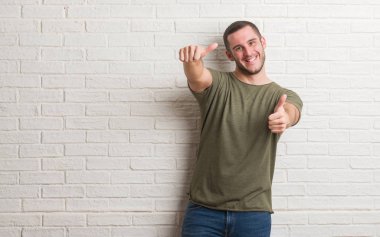 Genç beyaz adam ayakta beyaz tuğla duvar onaylama üzerinde el ile pozitif jest yapıyor gülümseyen ve başarı için mutlu yaşasın. Kamera, kazanan jest arıyorsunuz.