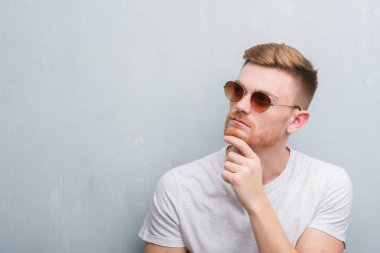 Gri grunge duvar retro güneş gözlüğü ciddi bir yüz düşünme hakkında soru, çok karışık fikir giyen genç Kızıl saçlı adam