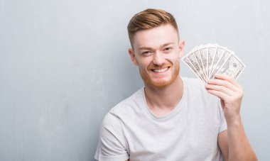 Genç Kızıl saçlı adam ayakta ve diş gösterilen kendine güvenen bir gülümsemeyle gülümseyerek mutlu bir yüz dolar tutan gri grunge duvarın üzerinden