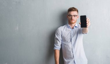 Gri grunge duvar gösteren boş ekran smartphone acımak yüz ciddi düşünüyorum üstünde kendine güvenen bir ifade ile genç Kızıl saçlı adam
