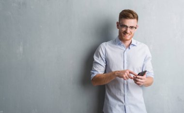 Genç Kızıl saçlı adam ayakta ve diş gösterilen kendine güvenen bir gülümsemeyle gülümseyerek mutlu bir yüz ile smartphone kullanarak ileti gönderme gri grunge duvarın üzerinden