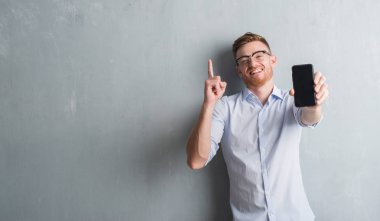 Genç Kızıl saçlı adam akıllı telefon boş ekran gösterilen gri grunge duvarın üzerinden bir fikir ya da soru işaret parmağı gülen yüz, bir numara ile şaşırttı