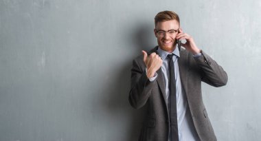 Genç redhead iş adamı işaret ve başparmak kadar mutlu yüz gülümseyen tarafı ile gösterilen telefonda konuşurken gri grunge duvarın üzerinden