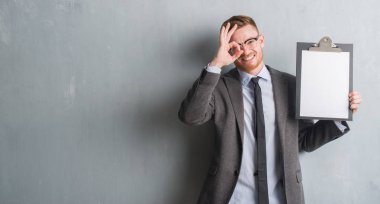 Genç redhead iş adamı Pano ile ok işareti ile el parmaklarının arasından seyir göze yapıyor gülümseyen mutlu yüz tutan gri grunge duvarın üzerinden