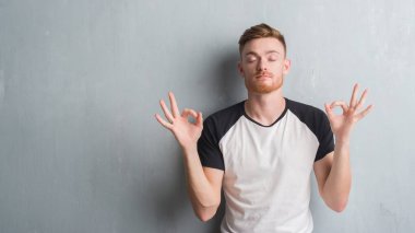Genç Kızıl saçlı adam gri grunge duvarın üzerinden rahatla ve gülümseyen gözlerle kapalı parmak jestle meditasyon yapıyor. Yoga kavramı.
