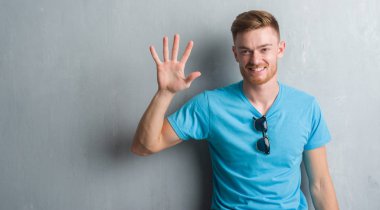 Young redhead man over grey grunge wall wearing casual outfit showing and pointing up with fingers number five while smiling confident and happy. clipart