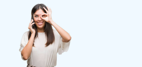 Young beautiful hispanic using smartphone with happy face smiling doing ok sign with hand on eye looking through fingers