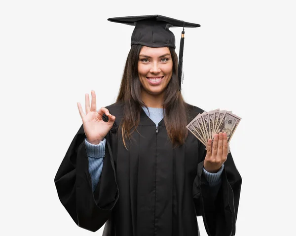 年轻的西班牙裔女子穿着毕业制服持有美元做 标志与手指 优秀的符号 — 图库照片
