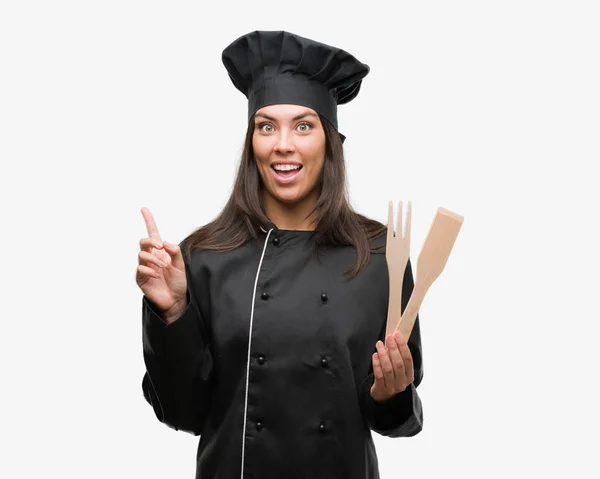 年轻的西班牙裔厨师妇女穿着厨师制服惊讶的想法或问题指向手指与快乐的脸 — 图库照片