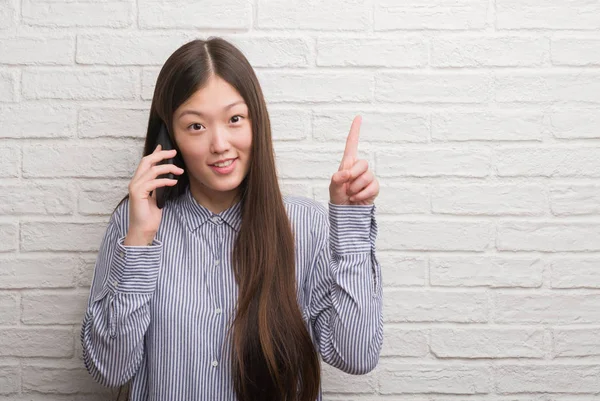 在墙上的年轻中国妇女谈论电话惊讶的想法或问题指着一个快乐的脸 — 图库照片