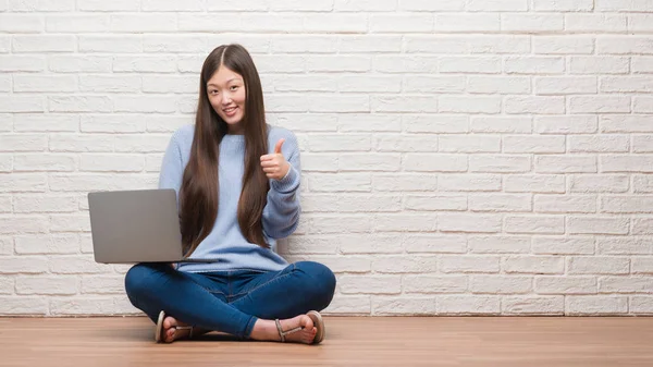 年轻的中国妇女坐在地板上墙上使用笔记本电脑快乐的大微笑做 拇指与手指 优秀的标志 — 图库照片