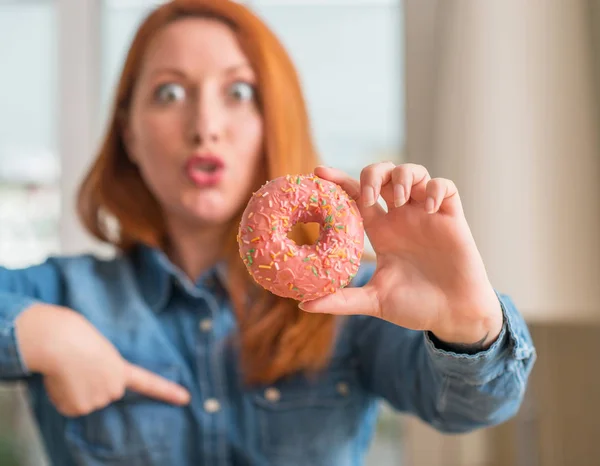 赤毛の女性が自分自身に驚き顔人差し指と家庭でドーナツを保持 — ストック写真