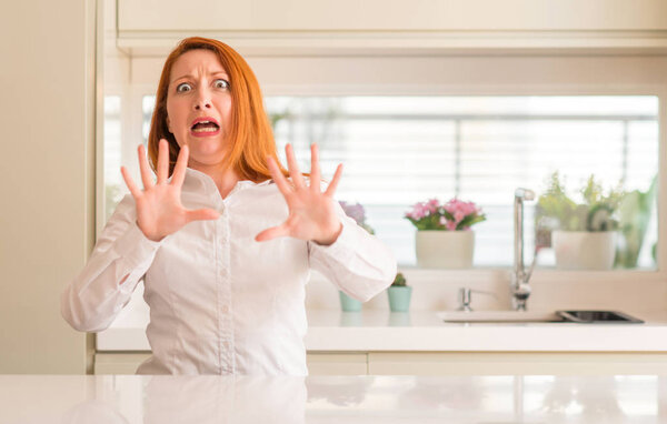 Redhead woman at kitchen afraid and terrified with fear expression stop gesture with hands, shouting in shock. Panic concept.