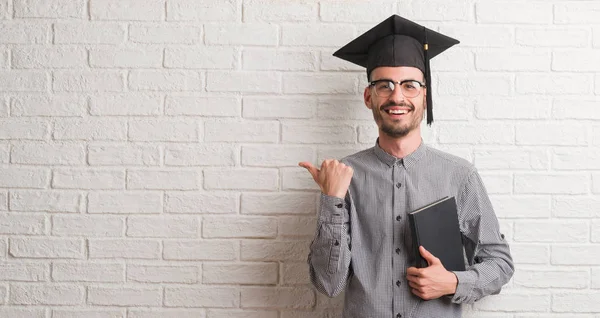 指していると幸せそうな顔を笑顔で側まで親指で示す卒業帽をかぶってレンガ壁を越えて若い成人男性 — ストック写真