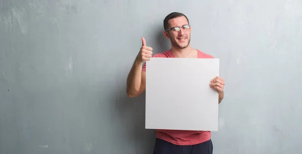 Homem Caucasiano Jovem Sobre Parede Grunge Cinza Segurando Banner Branco — Fotografia de Stock