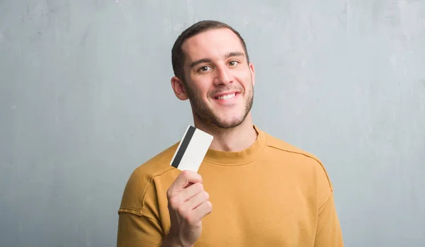 Young Caucasian Man Grey Grunge Wall Holding Credit Card Happy — Stock Photo, Image