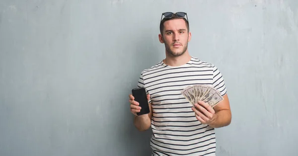 Young Caucasian Man Grey Grunge Wall Holding Dollars Smarphone Serious — Stock Photo, Image