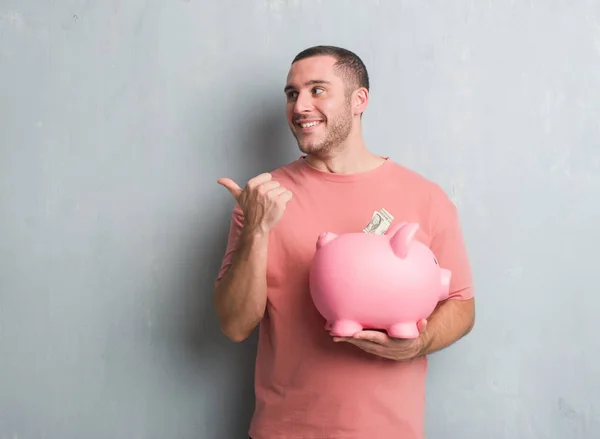 Young Caucasian Man Grey Grunge Wall Holding Piggy Bank Pointing — Stock Photo, Image