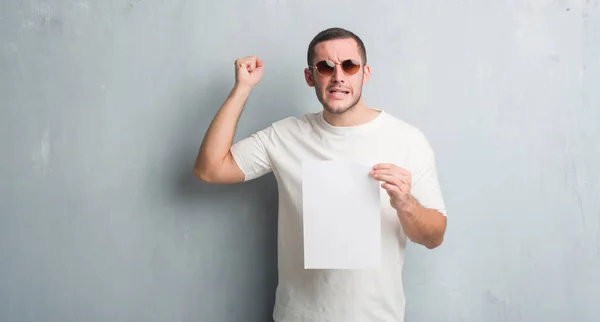 Jovem Caucasiano Sobre Parede Grunge Cinza Segurando Papel Branco Irritado — Fotografia de Stock