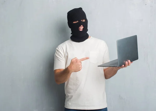 年轻的白种人黑客在灰色的垃圾墙做网络攻击使用膝上型电脑非常高兴地指向用手和手指 — 图库照片